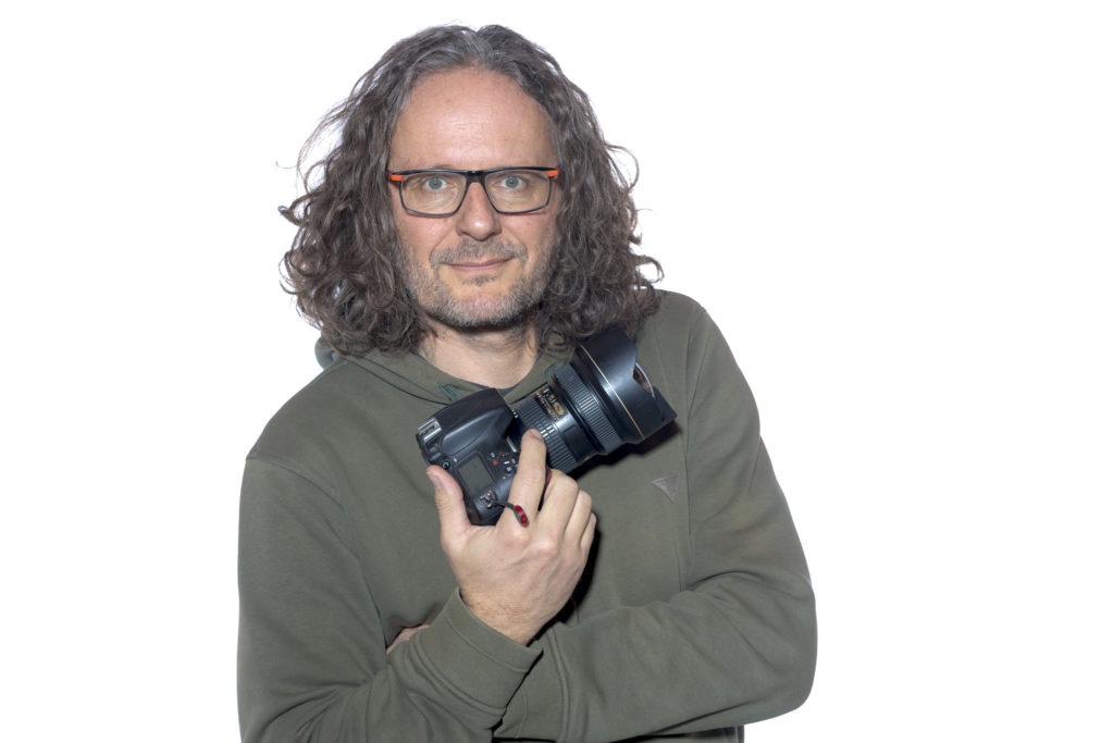 Autoportrait de David Casartelli sur fond blanc portant en main son appareil photo